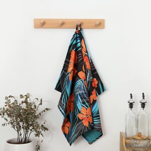 May include: A black kitchen towel with a vibrant floral pattern of orange hibiscus flowers and turquoise leaves. The towel is hanging from a wooden peg rack on a white wall. A potted plant and kitchen accessories are in the background.