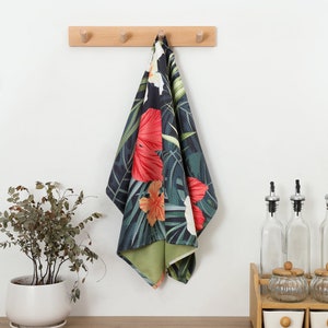 May include: A rectangular kitchen towel featuring a dark background with a vibrant tropical print of large red, orange, and white hibiscus flowers and green palm leaves. The towel has a textured surface and a solid light green underside.