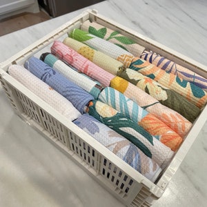 May include: A white plastic storage bin with a grid pattern holds a stack of colorful kitchen towels with tropical leaf and floral patterns.