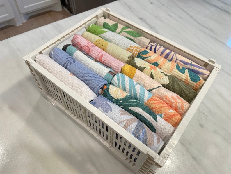 May include: A white plastic storage bin with a grid pattern holds a stack of colorful kitchen towels with tropical leaf and floral patterns.