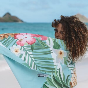 May include: A woman with curly hair is partially hidden behind a blue beach towel with a tropical floral print. The towel features hibiscus flowers, palm leaves, and pineapples.