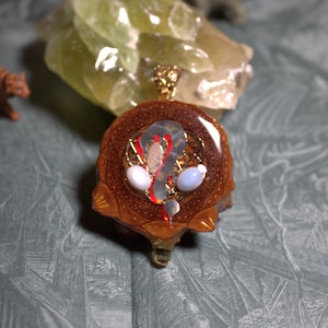 May include: A circular pendant featuring a brown, textured pine cone base with a glossy resin center. Embedded within the resin are gold wire coils, two smooth white oval stones, and thin red and iridescent accents, topped with a gold metal bail.