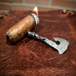 May include: A lit cigar with a metal cigar cutter on a brown leather surface. The cigar is burning at the tip, with a flame visible. The cutter is made of metal and has a unique, textured design.