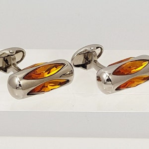 May include: A pair of silver cufflinks with amber-colored glass inserts. The glass is cut into a geometric shape and is set in a silver frame.