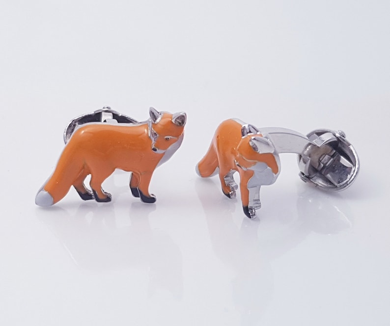 May include: A pair of silver cufflinks featuring a fox design. The foxes are orange with black eyes and noses. The cufflinks are a great way to add a touch of whimsy to any outfit.