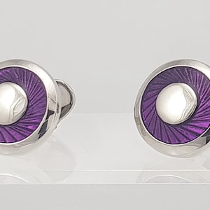 May include: A pair of silver cufflinks with a circular design. Each cufflink features a central, raised, silver-colored circle surrounded by a ring of radiating purple lines. The cufflinks are presented on a light background.