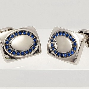 Könnte beinhalten: Ein Paar silberne Manschettenknöpfe mit einem blauen ovalen Design. Das Oval ist von einem Ring aus kleinen blauen Steinen umgeben.