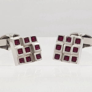 May include: A pair of silver cufflinks with a square design featuring a grid of red gemstones.