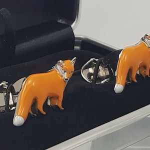 May include: A pair of silver cufflinks with orange enamel fox designs. The cufflinks are in a black velvet box.