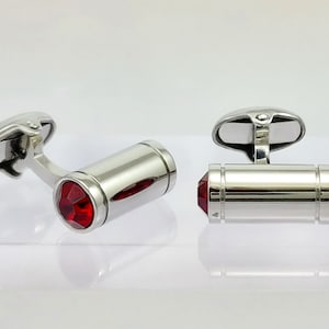 May include: A pair of silver cufflinks with a polished finish. Each cufflink features a cylindrical design with a vibrant red gemstone at each end. The cufflinks are displayed on a reflective surface.