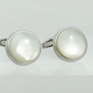 May include: A pair of silver cufflinks with a round, pearlescent face. The cufflinks have a polished silver border with a subtle grooved design. The background is a bright, neutral color.