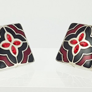 Peut inclure: Une paire de boutons de manchette carrés argentés arborant un motif décoratif avec des sections en émail noir et rouge foncé. Le centre de chaque bouton présente une fleur à quatre pétales ornée de petits cristaux rouges facettés.