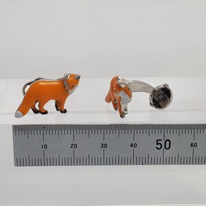 May include: A pair of silver fox cufflinks with orange enamel. The cufflinks are shown on a white surface with a ruler for scale.