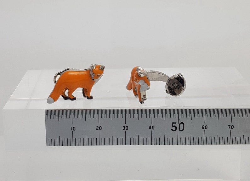 May include: A pair of silver fox cufflinks with orange enamel. The cufflinks are shown on a white surface with a ruler for scale.