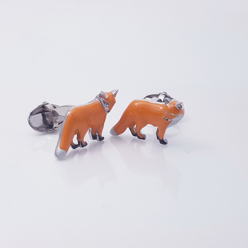 May include: A pair of silver fox cufflinks with orange enamel. The foxes are facing forward with their tails curled behind them.