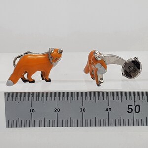 May include: A pair of silver fox cufflinks with orange enamel. The cufflinks are shown next to a ruler, measuring approximately 1.97 inches in length.