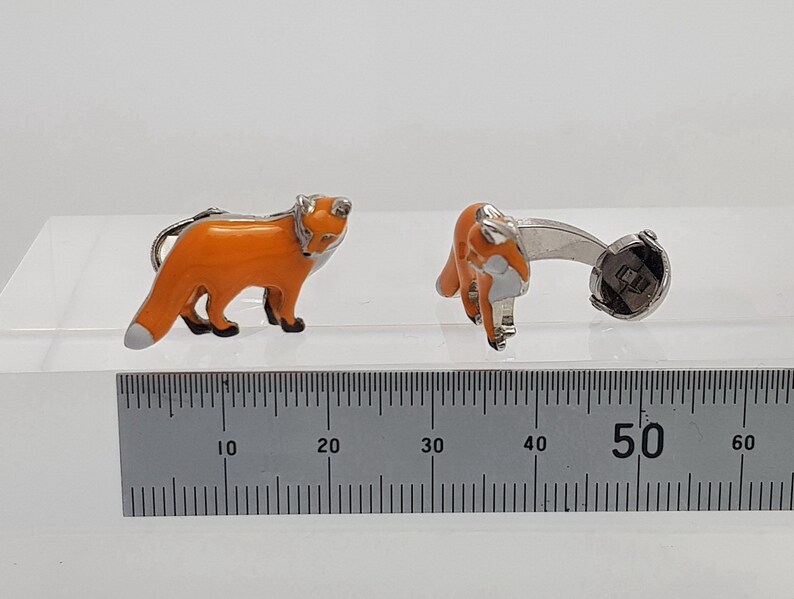May include: A pair of silver fox cufflinks with orange enamel. The cufflinks are shown next to a ruler, measuring approximately 1.97 inches in length.