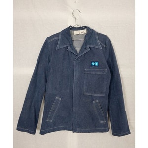 Puede incluir: Una chaqueta vaquera azul oscuro con cuello blanco y un pequeño parche en el bolsillo. El parche es azul y blanco.