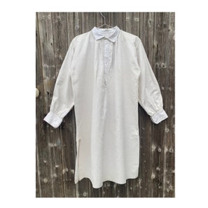 May include: A white long-sleeved shirt with a button-up collar and a long, flowing design. The shirt has a single button closure at the neck and a split hem.