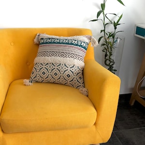 Op de afbeelding: Een decoratief kussen met een geometrisch patroon in teal, crème en bordeauxrood ligt op een gele fauteuil. Het kussen heeft aan elke hoek kwastjes. Op de achtergrond staat een hoge glazen vaas met een plant.