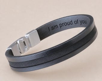 May include: A black leather bracelet featuring a silver-toned metal clasp and the engraved text "I am proud of you" on the inner surface. The bracelet consists of two parallel bands joined together, creating a layered appearance with a smooth finish.