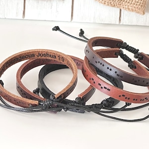 May include: A collection of leather bracelets in various shades of brown and black. Some bracelets feature engraved text, while others display a Morse code pattern. Each bracelet has an adjustable black cord closure.