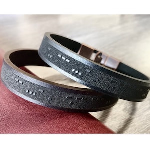 Pode incluir: Duas pulseiras de couro preto com letras em braille em relevo. As pulseiras estão empilhadas uma em cima da outra.