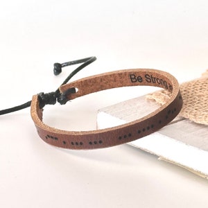 May include: Brown leather bracelet with black stitching and the words "Be Strong" engraved on the front. The bracelet is tied with a black cord.