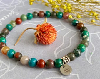 Puede incluir: Una pulsera de cuentas con una variedad de piedras preciosas de colores, incluyendo turquesa, rojo y verde. La pulsera tiene un dije plateado con un diseño floral. Una flor naranja y ramitas amarillas en el fondo.