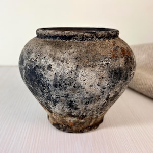 May include: A small, round, weathered ceramic pot with a wide opening and a thick, dark rim. The exterior surface features a mottled, textured finish in shades of charcoal grey, black, and muted brown, showing signs of age and wear throughout.