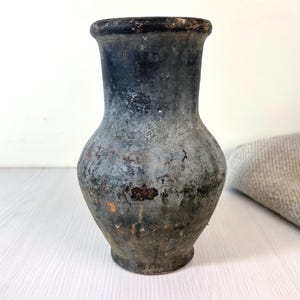 May include: A dark, weathered ceramic vase with a bulbous body and a flared rim. The surface features a mottled, uneven finish with shades of charcoal, black, and hints of rust-coloured brown, showing signs of age and a textured, matte appearance.