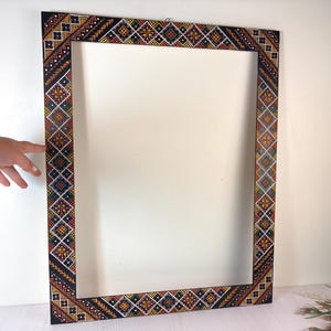 Könnte beinhalten: Ein rechteckiger Bilderrahmen mit schwarzem Untergrund und handgemalten geometrischen Mustern in Gelb, Orange und Weiß. Das Design besteht aus sich wiederholenden Rautenformen, kleinen Punkten und linearen Zickzack-Akzenten am Rand.