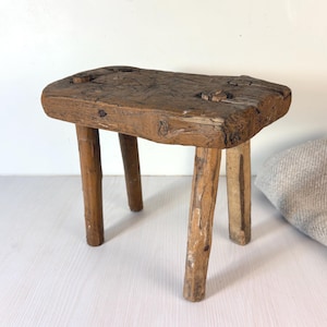 Puede incluir: Un pequeño taburete de madera rústica con una tapa rectangular gruesa con uniones visibles y cuatro patas cilíndricas robustas. La madera muestra un acabado marrón oscuro desgastado con vetas naturales, nudos y signos de envejecimiento.