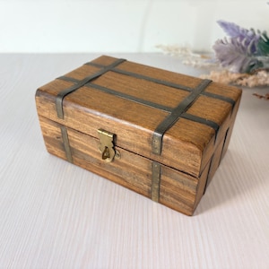 Puede incluir: Una caja rectangular de madera con un acabado de veta natural en color marrón, que presenta bandas metálicas oscuras sobre la tapa y los laterales. La caja incluye un cierre metálico de color latón en el centro para asegurar la tapa.
