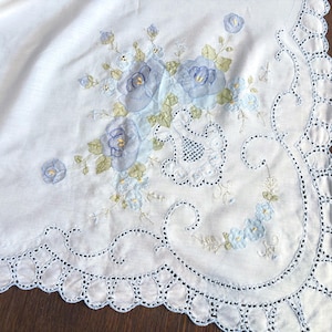 May include: A white fabric cloth featuring intricate blue and green floral embroidery. The design includes large blue roses, smaller blossoms, and green leaves. The edges are finished with a decorative scalloped border and cutwork detailing.