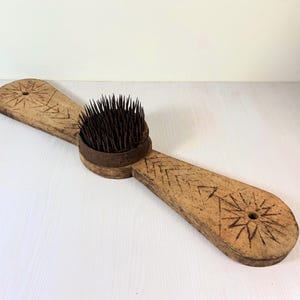 May include: A long, light brown wooden tool featuring a central circular metal base filled with numerous upright, dark, thin metal spikes. The wooden handle extends on both sides, decorated with carved starburst patterns and chevron lines.