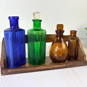 May include: A collection of four vintage glass bottles in blue, green, and amber hues, displayed on a small wooden shelf. The blue and green bottles feature embossed text reading NOT TO BE TAKEN, while the amber bottles have rounded and rectangular shapes.