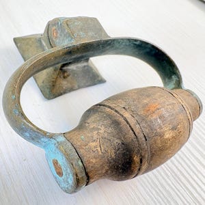 以下が含まれることがあります： 木製のハンドルと金属製のリングが付いたアンティークのドアノッカー。金属は、青と緑の緑青のヒントを持つ、風化した、経年変化した外観をしています。木製のハンドルには摩耗の兆候が見られます。