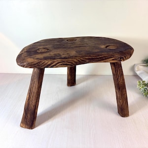 Può includere: Uno sgabello piccolo in legno a tre gambe con una finitura tinta in marrone scuro. La seduta ha una forma arrotondata irregolare con venature del legno visibili e tre rientranze circolari sulla superficie superiore, sostenuto da tre gambe angolate.
