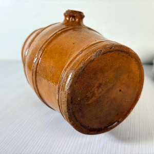 Pode incluir: Uma jarra cilíndrica de grés castanho com um esmalte brilhante e faixas horizontais em relevo que rodeiam o corpo. O recipiente apresenta um pequeno bico centrado no topo e uma base circular plana, com uma estética rústica e terrosa.