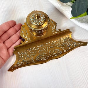 Puede incluir: Tintero de latón dorado ornamentado. Presenta un tintero redondo con tapa abovedada y una base rectangular con intrincados diseños florales. El latón tiene un cálido tono dorado con detalles blancos.