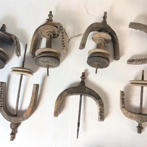 May include: A collection of seven weathered wooden spinning wheel flyers and bobbins. These antique components feature U-shaped wooden frames with small metal hooks, central spindles, and circular wooden whorls, showing natural wood grain and age.