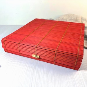May include: A rectangular box constructed from thin, red-dyed bamboo slats held together by brown thread. The box features a hinged lid and a small, light-coloured wooden toggle closure centred on the front edge, showcasing a slatted texture.
