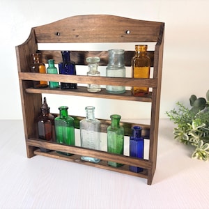 May include: A dark brown wooden wall-mounted shelf with two tiers holding eleven vintage glass bottles in various colours including amber, cobalt blue, emerald green, and clear glass. The shelf features a curved top edge and two mounting holes.
