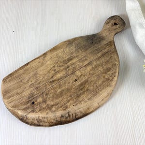 Puede incluir: Una tabla de cortar rústica de madera con un asa. La tabla es de color marrón claro con vetas de madera visibles y un aspecto ligeramente desgastado. El asa tiene un agujero para colgar. Un paño blanco y vegetación en el fondo.
