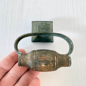 May include: Antique bronze drawer pull with a square backplate and an oval handle. The handle has a cylindrical center section with vertical grooves. The metal has a weathered, aged appearance.