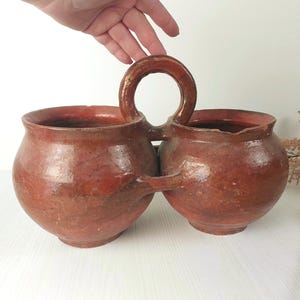 Puede incluir: Un recipiente de terracota de doble cámara con un acabado vidriado de color marrón rojizo, dos cuencos redondeados conectados por un asa central arqueada y un soporte tipo puente entre las bases, con una construcción de arcilla rústica.