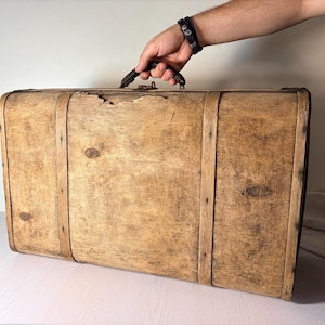 May include: A rectangular wooden suitcase with a natural light brown wood grain finish, featuring two vertical wooden support straps, a dark handle, and visible wear including a small chip on the top edge near the centre latch mechanism.