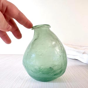 Puede incluir: Una botella de vidrio verde claro y texturizado con un cuerpo redondeado y un cuello estrecho. La botella tiene una forma ligeramente irregular, lo que sugiere que está hecha a mano. La botella está vacía y parece limpia.