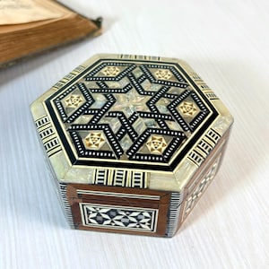 May include: A decorative, hexagonal wooden box with intricate inlaid mother-of-pearl and geometric patterns. The box features a star design on the lid, with black and gold accents. The box is a vintage item.